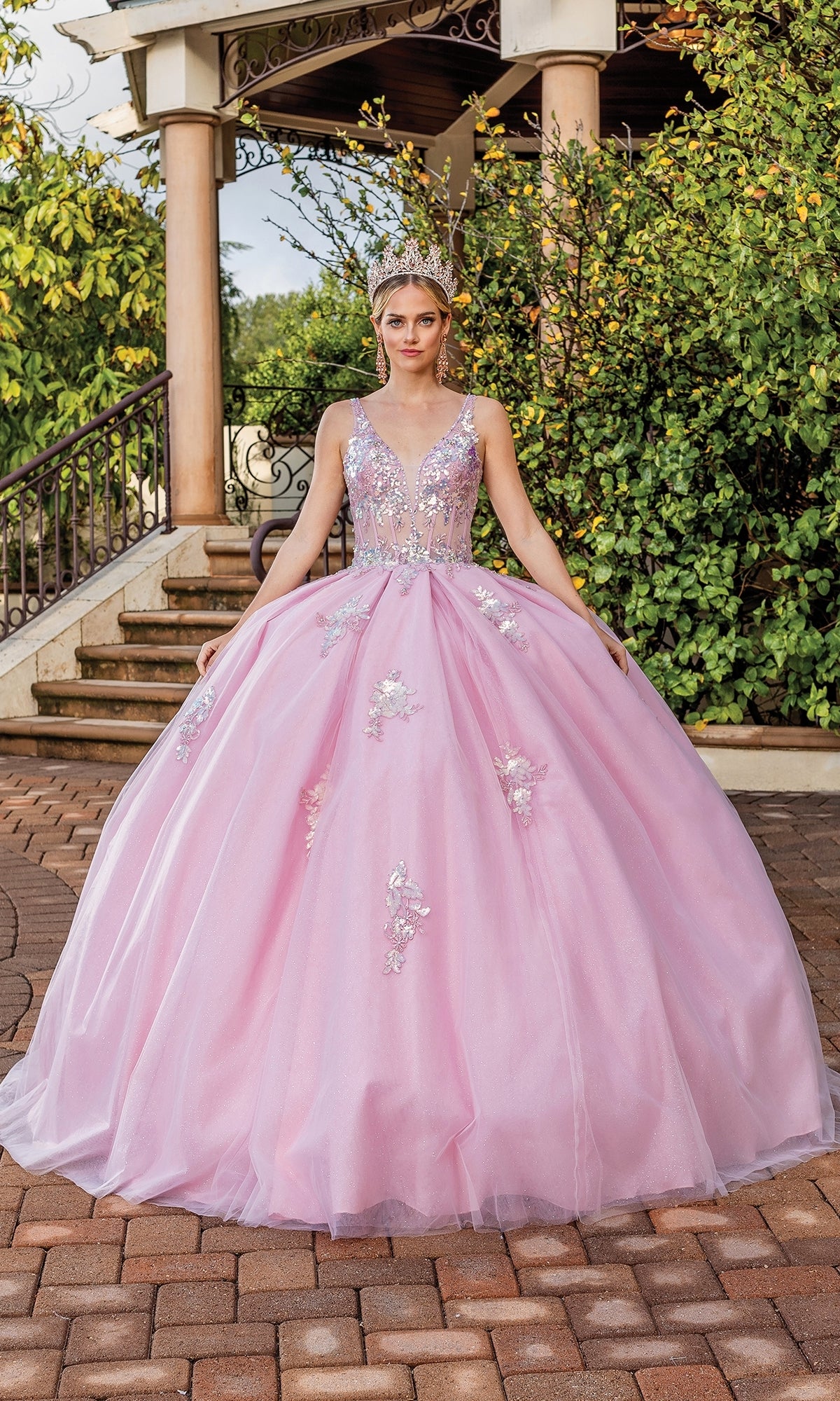 Wearing the Quinceanera Dress 1896 By Dancing Queen, a woman stands outdoors on a brick path before steps and greenery, stunning in a blush pink gown with silver embellishments, lace embroidery, and a tiara.